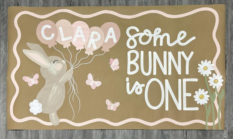 May include: A rectangular tan sign featuring a painted illustration of a light brown bunny holding five pink balloons with the name CLARA written on them. The text reads SOME BUNNY is ONE, accompanied by three small pink butterflies and white daisies.