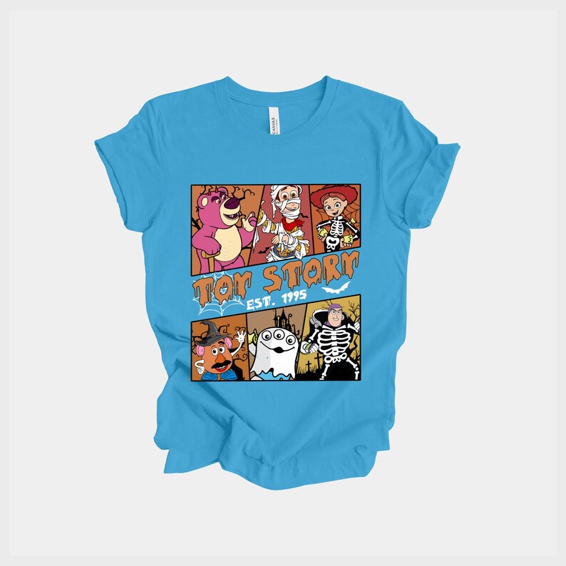 K&ouml;nnte beinhalten: Hellblaues T-Shirt mit einem Design aus dem Film Toy Story. Das Design zeigt Charaktere in Halloween-Kost&uuml;men, mit dem Text "Toy Story Est. 1995" in Orange. Das T-Shirt ist aus weichem Material.