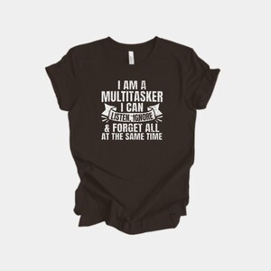 Puede incluir: Una camiseta marrón oscuro con el texto blanco: "I AM A MULTITASKER I CAN LISTEN, IGNORE & FORGET ALL AT THE SAME TIME". El texto está dispuesto en formato apilado con un diseño de banner.