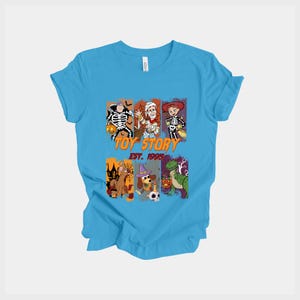 May include: A light blue t-shirt featuring a graphic of Toy Story characters dressed in Halloween costumes. The graphic includes the text "TOY STORY EST. 1995". The design features characters like Woody, Buzz, and Rex in Halloween attire.