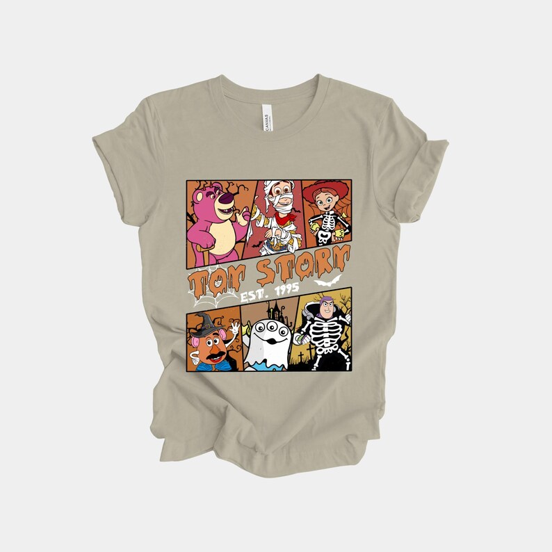 K&ouml;nnte beinhalten: Beigefarbenes T-Shirt mit einem farbenfrohen Motiv von Toy Story-Charakteren in Halloween-Kost&uuml;men. Das Design zeigt Lotso, Woody, Buzz Lightyear, Mr. Potato Head, einen Geist und ein Skelett. Der Text "Toy Story Est. 1995" ist ebenfalls zu sehen.