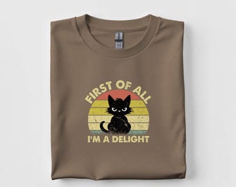 May include: Brown t-shirt with a graphic of a black cat inside a retro sunset design. The text reads "FIRST OF ALL I'M A DELIGHT" in a distressed font. The shirt is folded.