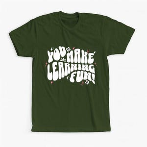 Op de afbeelding: Donkergroen t-shirt met de witte tekst "YOU MAKE LEARNING FUN!" in een gestileerd lettertype. Het ontwerp bevat kleine bloem- en steraccenten in roze. Het t-shirt is effen van kleur en heeft een ronde hals.
