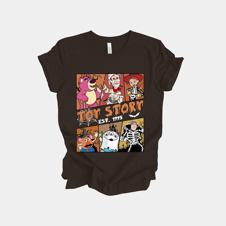 K&ouml;nnte beinhalten: Ein dunkelbraunes T-Shirt mit einer farbenfrohen Grafik von Toy Story-Charakteren in Halloween-Kost&uuml;men. Das Design umfasst Lotso, Woody und andere, mit dem Text "Toy Story Est. 1995". Das Gesamtthema ist eine Mischung aus dem beliebten Film und Halloween.