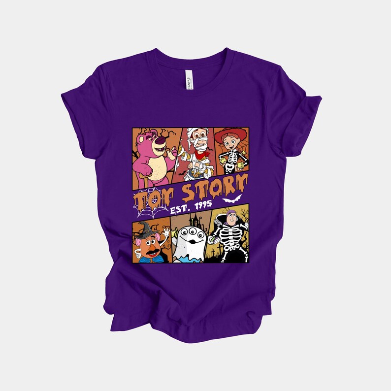 K&ouml;nnte beinhalten: Ein lila T-Shirt mit einer Grafik von Toy Story-Charakteren in Halloween-Kost&uuml;men. Das Design umfasst Lotso, Woody, Jessie, Mr. Potato Head, ein Gespenst und ein Skelett. Der Text "Toy Story Est. 1995" ist ebenfalls aufgedruckt.