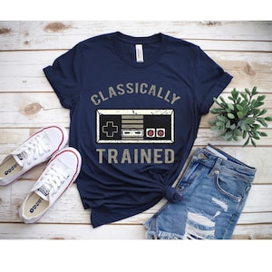 May include: Navy blue t-shirt with the text "CLASSICALLY TRAINED" and a graphic of a retro video game controller. The shirt is paired with white sneakers and blue denim shorts, creating a casual, everyday look.