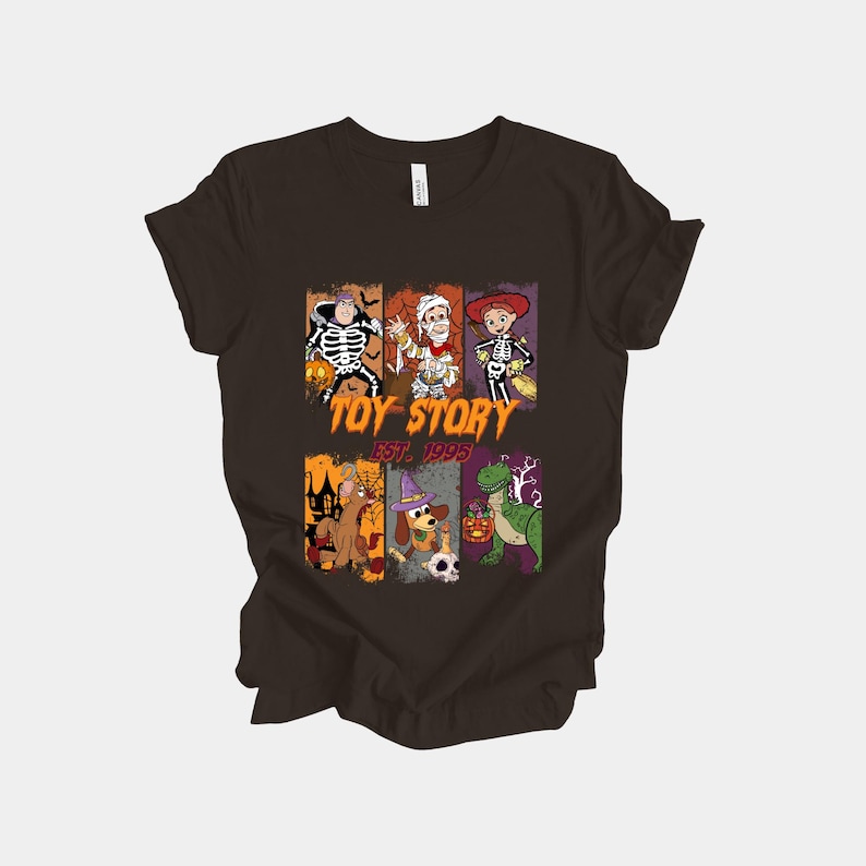 May include: Dark brown t-shirt featuring a Halloween-themed graphic with Toy Story characters dressed in costumes. The graphic includes Buzz Lightyear as a skeleton, Woody as a mummy, and Jessie as a skeleton. The text "TOY STORY EST. 1995" is also included.