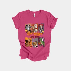 May include: A magenta t-shirt featuring a graphic design with characters from Toy Story dressed in Halloween costumes. The design includes the text "TOY STORY EST. 1995".