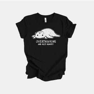 May include: Black t-shirt with a white illustration of a seal-like animal. The text "OVERTHINKING AND ALSO HUNGRY" is printed below the illustration. The shirt is made of a soft material and is suitable for casual wear.