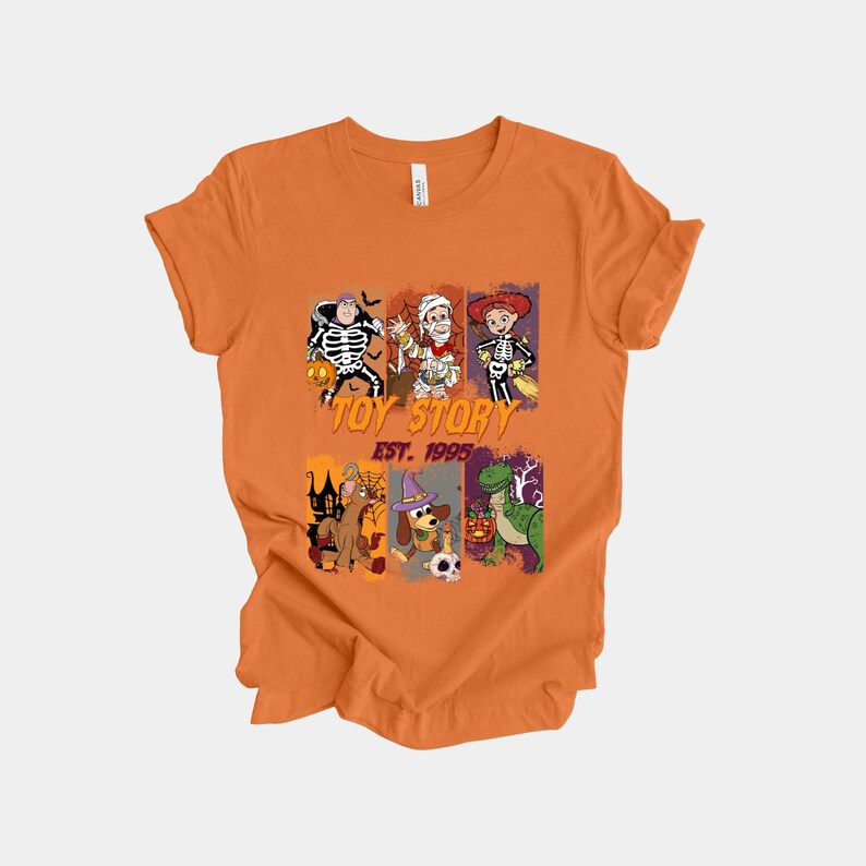 May include: Orange t-shirt featuring a graphic design with characters from Toy Story dressed in Halloween costumes. The text "TOY STORY EST. 1995" is printed on the shirt. The design includes images of Woody, Buzz Lightyear, and other characters.