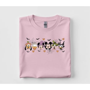 Puede incluir: Una camiseta rosa claro con un diseño temático de Halloween. El diseño incluye personajes de dibujos animados disfrazados para Halloween, con murciélagos, telarañas y calabazas. La camiseta está doblada.