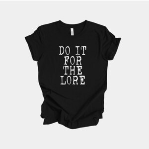 Może przedstawiać: Czarny t-shirt z białym napisem "DO IT FOR THE LORE" pogrubioną czcionką w stylu vintage. Koszulka wykonana jest z miękkiego materiału i ma klasyczny okrągły dekolt. Casualowy i wygodny element garderoby.