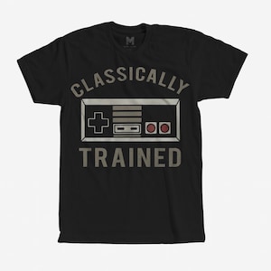 May include: Black t-shirt with the text "CLASSICALLY TRAINED" and a graphic of a retro video game controller in white and gray. The shirt has a crew neck and short sleeves.
