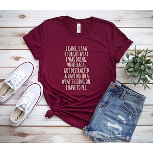 Pode incluir: Camiseta bordô com texto branco: "I CAME, I SAW I FORGOT WHAT I WAS DOING. WENT BACK, GOT DISTRACTED & HAVE NO IDEA WHAT'S GOING ON. I HAVE TO PEE." Combinada com shorts jeans e tênis brancos.