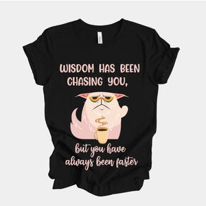 Puede incluir: Camiseta negra con un gráfico de un gato blanco gruñón sosteniendo una taza de café. El texto rosa dice "WISDOM HAS BEEN CHASING YOU, but you have always been faster". Un diseño divertido para uso casual.