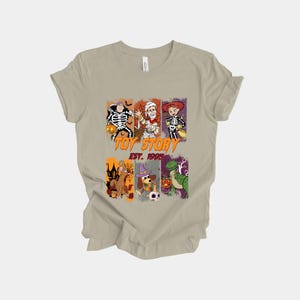 May include: A tan t-shirt featuring a graphic design with characters from Toy Story in Halloween costumes. The design includes the text "TOY STORY EST. 1995" in orange and green. The shirt is made of a soft material.