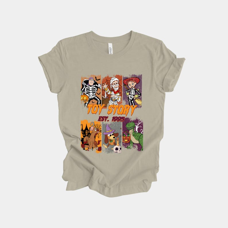 May include: A tan t-shirt featuring a graphic design with characters from Toy Story in Halloween costumes. The design includes the text "TOY STORY EST. 1995" in orange and green. The shirt is made of a soft material.