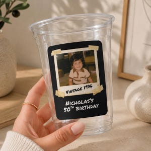May include: A clear plastic disposable cup featuring a rectangular black label with a vintage-style photo of a child. The label includes the text "VINTAGE 1976" and "NICHOLAS'S 50TH BIRTHDAY" printed in white, with faux tape accents on the photo corners.