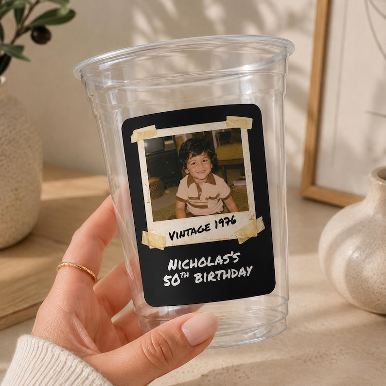 May include: A clear plastic disposable cup featuring a rectangular black label with a vintage-style photo of a child. The label includes the text "VINTAGE 1976" and "NICHOLAS'S 50TH BIRTHDAY" printed in white, with faux tape accents on the photo corners.