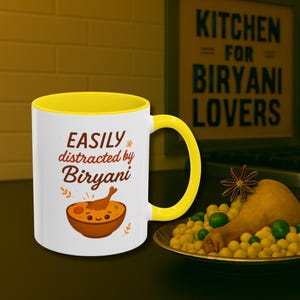 Könnte beinhalten: Weißer Keramikbecher mit gelbem Innenraum und Henkel. Der Becher hat den Text "EASILY distracted by Biryani" und eine Illustration einer Biryani-Schüssel. Im Hintergrund befindet sich ein Teller mit Biryani.