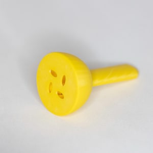 May include: A bright yellow cheese slicer with a handle. The slicer has a rounded head with four oval-shaped holes. The handle is cylindrical and has a ridged texture. The slicer is made of plastic and is on a white background.