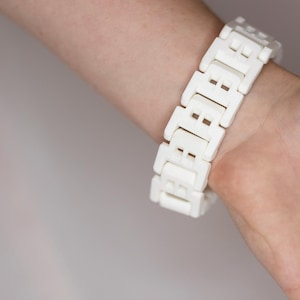 May include: A white, rectangular link bracelet is worn on a wrist. The bracelet is made of multiple, identical links, each with a rectangular shape and a small, square cutout. The bracelet is approximately 7 inches in length.
