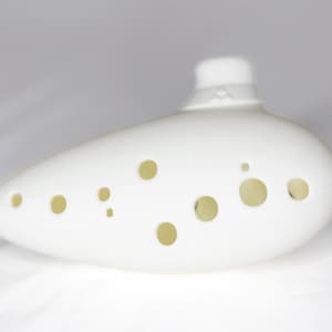 May include: A white ceramic ocarina, a wind instrument, with a rounded body and a mouthpiece. The ocarina has several finger holes of varying sizes. The instrument is set against a white background.