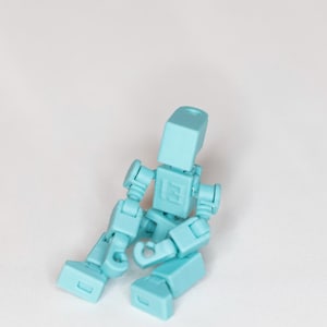 May include: A light blue articulated action figure in a seated position. The figure has a blocky design with multiple joints, allowing for various poses. The figure is made of plastic and is resting on a white surface.