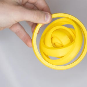 May include: A bright yellow, abstract 3D-printed sculpture with a circular outer frame and interwoven internal shapes. The sculpture is held by a hand against a neutral gray background. The design is modern and geometric.