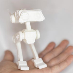 Könnte beinhalten: Ein kleines, weißes, bewegliches Roboterspielzeug. Es hat einen rechteckigen Kopf mit zwei kreisförmigen augenähnlichen Merkmalen und eine Reihe von rechteckigen und zylindrischen Komponenten. Das Spielzeug wird in einer offenen Hand vor weißem Hintergrund gehalten.