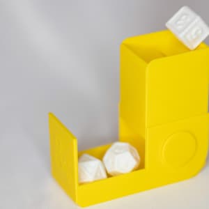 Op de afbeelding: Felgele gele dobbelsteentoren met twee witte dobbelstenen binnenin en één bovenop. De dobbelsteentoren is een container voor het gooien van dobbelstenen, vaak gebruikt in tabletop games. De dobbelstenen zijn wit met zwarte cijfers.