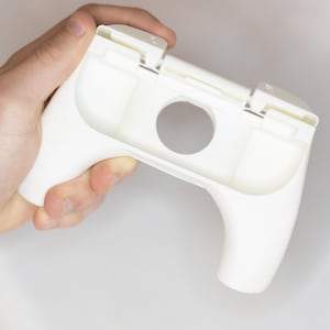 May include: A white plastic game controller with a rounded handle and a central circular opening. The controller has a rectangular top section with two raised areas. The design is simple and modern.