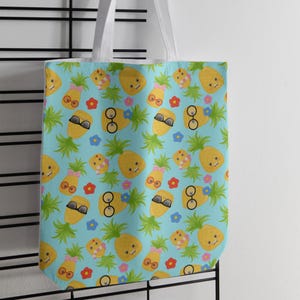 May include: A light blue tote bag featuring a repeating pattern of yellow pineapples with green leaves. The pineapples have various expressions and accessories, including pink bows, round glasses, and black sunglasses, interspersed with small red and blue flowers.