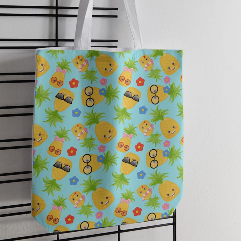 May include: A light blue tote bag featuring a repeating pattern of yellow pineapples with green leaves. The pineapples have various expressions and accessories, including pink bows, round glasses, and black sunglasses, interspersed with small red and blue flowers.