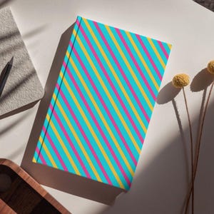 May include: A rectangular hardcover notebook featuring a vibrant pattern of diagonal stripes in alternating colors of bright turquoise, hot pink, and golden yellow. The notebook is positioned at an angle, showcasing its clean, flat front cover design.