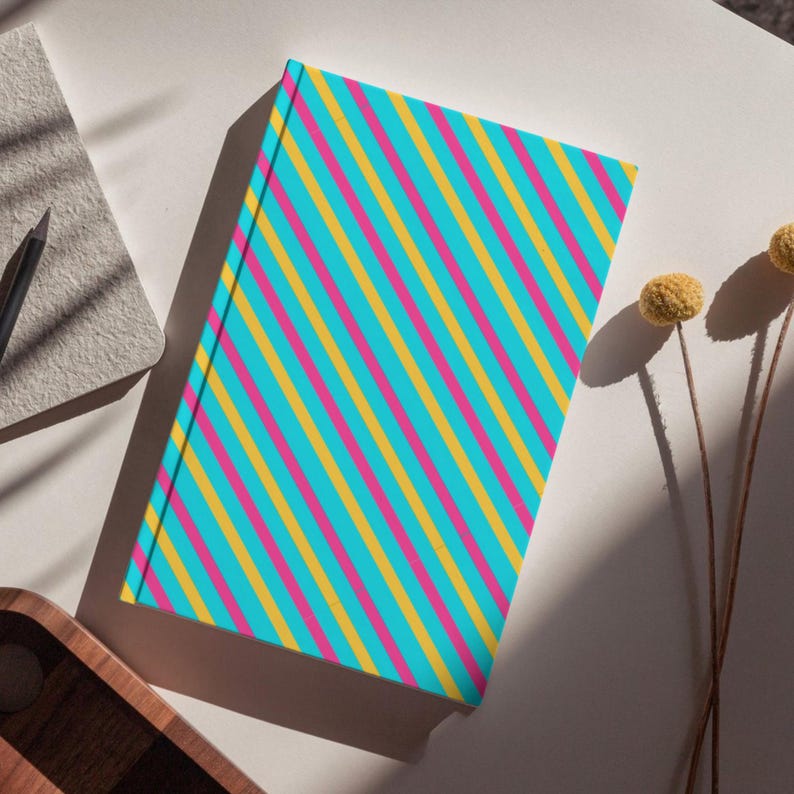 May include: A rectangular hardcover notebook featuring a vibrant pattern of diagonal stripes in alternating colors of bright turquoise, hot pink, and golden yellow. The notebook is positioned at an angle, showcasing its clean, flat front cover design.