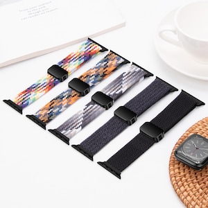 Puede incluir: Cinco correas de reloj tejidas de colores, con conectores negros. Las correas presentan una variedad de patrones y colores, incluyendo arcoíris, naranja, gris y negro. Un smartwatch es visible en el fondo.