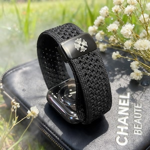 Puede incluir: Correa negra para Apple Watch con diseño tejido. La hebilla de metal negro presenta un diseño floral blanco. El reloj descansa sobre una superficie de cuero negro. Un accesorio elegante.