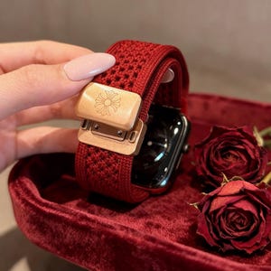 Könnte beinhalten: Ein rotes Uhrenarmband mit gewebter Textur und einem goldfarbenen Verschluss. Der Verschluss hat ein Blumenmuster. Das Armband wird auf einer roten Samtoberfläche mit zwei roten Rosen präsentiert.