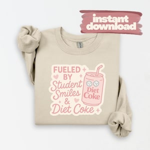 Op de afbeelding: Een beige sweatshirt met een roze afbeelding met de tekst "Fueled by Student Smiles & Diet Coke". De afbeelding bevat een illustratie van een Diet Coke-blikje en een roze banner met "instant download".