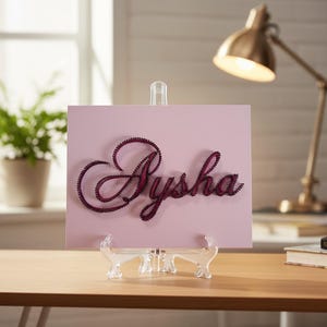 Könnte beinhalten: Ein rosa Schild mit dem Namen "Aysha" in dunkelroter String-Art-Technik. Das Schild wird auf einer transparenten Kunststoffstaffelei präsentiert. Eine goldene Schreibtischlampe und eine Topfpflanze sind im Hintergrund zu sehen.