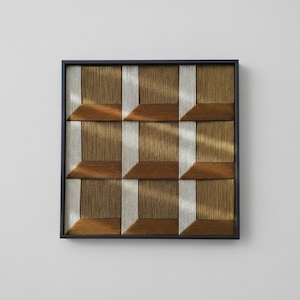 Op de afbeelding: Een vierkant wandkunstwerk met een geometrisch patroon van geweven draden in metallic goud-, brons- en zilverkleuren. Het ontwerp bestaat uit een raster van negen vierkanten in een dunne zwarte lijst, wat een driedimensionaal effect creëert.