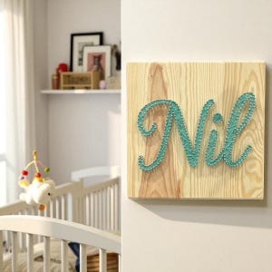 Puede incluir: Un letrero cuadrado de madera con el nombre "Nil" en arte de hilo turquesa. El letrero está montado en una pared blanca, con una cuna y un móvil visibles en el fondo. La madera tiene una veta natural.