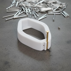 Puede incluir: Una herramienta blanca de forma ovalada con un clavo de latón insertado. Una pila de clavos plateados está dispersa en el fondo. La herramienta parece estar hecha de plástico y está diseñada para sujetar el clavo en su lugar.