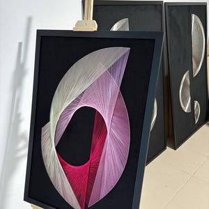 Pode incluir: Arte abstrata em fio sobre veludo preto, emoldurada em preto. A obra de arte apresenta um design de linhas que se cruzam em tons de branco, rosa e vermelho. A peça é exibida em um cavalete de madeira, com outras obras de arte emolduradas semelhantes ao fundo.