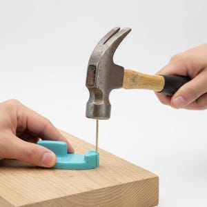 May include: A hammer strikes a nail into a wooden surface. A light blue tool is used to hold the nail in place. The hammer has a wooden handle and a black grip. The nail is brass-colored.