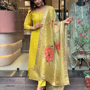 Op de afbeelding: Een heldergele salwar kameez-ensemble. De outfit bestaat uit een tuniek met lange mouwen, een bijpassende broek en een doorschijnende dupatta. De dupatta heeft een bloemenprint met roze en rode bloemen. De outfit is afgewerkt met gouden details.
