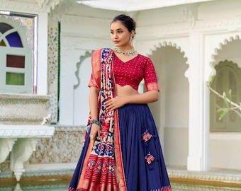 May include: A woman wearing a traditional Indian outfit. The ensemble includes a red blouse with short sleeves, a navy blue skirt, and a patterned shawl. The skirt and shawl feature intricate designs in red, blue, and gold. The woman is also wearing jewelry.