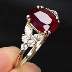 May include: A gold ring featuring a large, oval-cut ruby gemstone. The ruby is a deep red color and is surrounded by smaller, clear gemstones. The ring has a delicate, ornate design with additional small gemstones on the band.