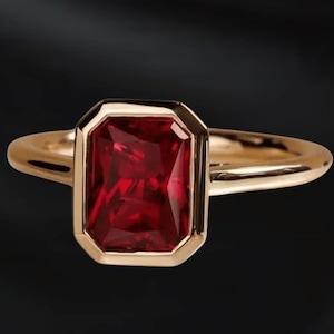 May include: A gold ring featuring a rectangular, deep red gemstone. The gemstone is set within a gold bezel, and the band is a simple, polished gold. The ring is displayed against a dark background.